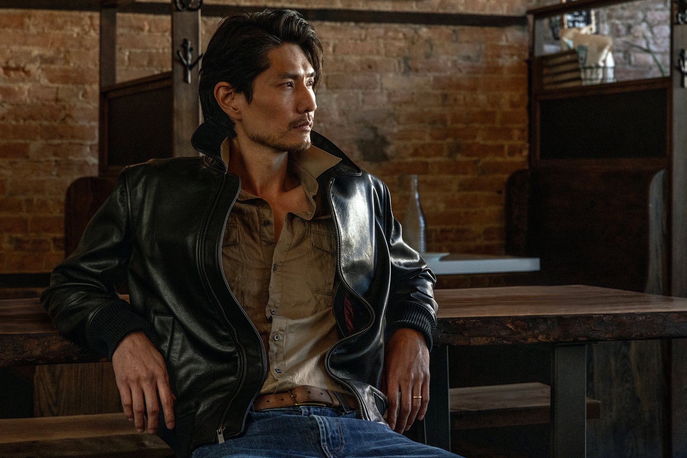 Man wearing a leather jacket sitting in a rustic setting with brick walls.