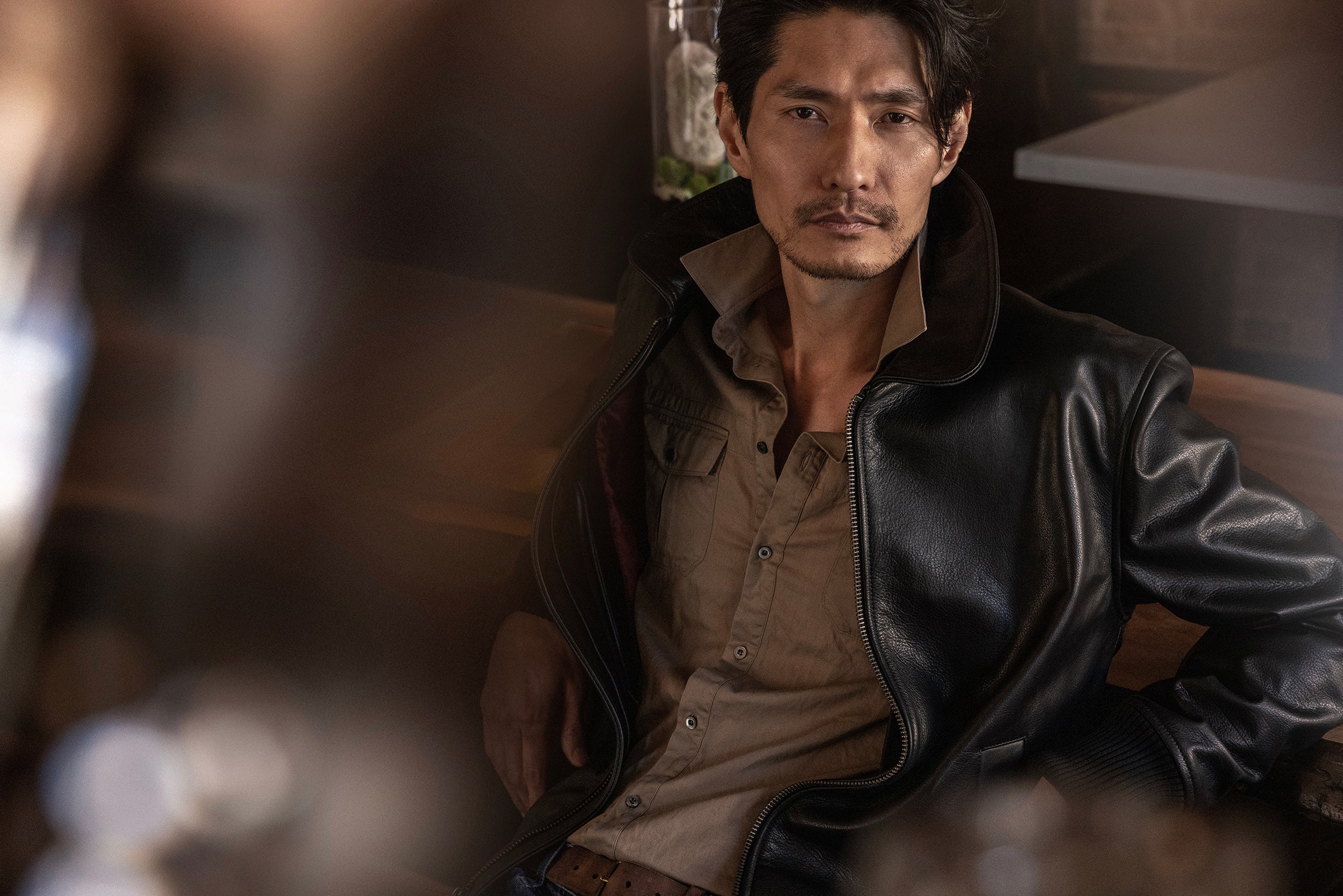 Man wearing a leather jacket and shirt sitting indoors with a blurred background
